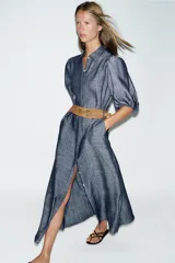 Vestido midi de denim azul con cuello camisero, mangas abullonadas y cierre frontal oculto con botones. Incluye un cinturón marrón con hebilla metálica.