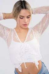Blusa de encaje floral blanco con transparencias, mangas largas, escote cruzado y lazos frontales con volados.
