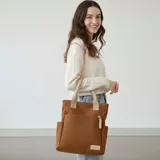 Cartera tipo tote de lona color marrón con asas de mano en color beige. Cuenta con un bolsillo frontal, bolsillos laterales y un detalle de tirador de cierre en cordón blanco.