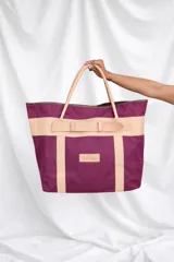 Bolso de playa grande confeccionado en lona color bordó, con detalles de asas y tiras frontales de cuero en tono tostado. Cuenta con un diseño estructurado y una etiqueta de cuero con la marca grabada en la parte frontal.