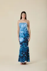 Vestido largo de corte sirena con escote recto y tirantes finos. Presenta un estampado floral en tonos azules sobre fondo claro, con una silueta ajustada que se ensancha suavemente hacia el ruedo.