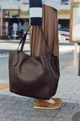 Bolso tipo shopper de gran tamaño, color marrón oscuro, con acabado brillante y doble asa de hombro con pespuntes decorativos. Cuenta con cierre mediante botón imantado y bolsillo interior con cremallera.