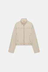 Chaqueta de cuello subido y manga larga en color beige con efecto lavado. Presenta cierre frontal con cremallera y detalle de tachas metálicas en forma de estrella distribuidas en el frente.