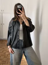 Sobrecamisa oversize de ecocuero negro, con cuello camisero, cierre frontal de botones, bolsillo en el pecho y mangas largas.