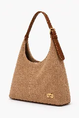 Cartera tipo hobo de gamuza color avena con diseño amplio y costuras decorativas. Cuenta con correa ajustable de cuero marrón y una placa metálica dorada con el logo de la marca en la parte inferior.
