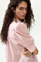 Blusa color rosado con cuello mao y botones ocultos en la parte delantera. Tiene canesú de doble capa con frunces adelante y atrás, mangas cortas abullonadas y puños con abertura y botón.