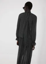 Blusa de poliéster color negro con estampado de lunares blancos, cuello alto con detalle de lazo, manga larga con puños elásticos y diseño recto.