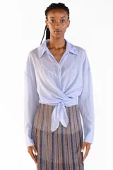 Camisa de manga larga color celeste, confeccionada en algodón, con cuello clásico, cierre frontal de botones y diseño cruzado con lazo para anudar en la cintura.