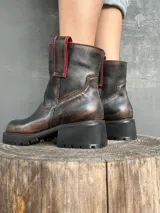 Bota de cuero con efecto desgastado en tonos marrones y negros, con suela track robusta y cierre lateral. Presenta detalles en color rojo en la parte superior de la caña.