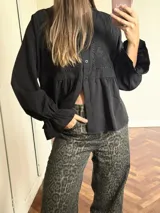 Camisa negra de corte peplum con detalles de bordado inglés en el pecho, cuello redondo y mangas largas con puños acampanados.