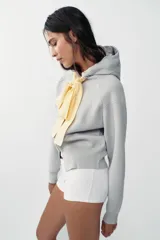 Chaqueta de punto con capucha, manga larga y cierre frontal con cremallera. Presenta un diseño de lazo decorativo en color amarillo en la zona del cuello.