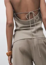 Top color beige, con cuello drapeado, diseño sin mangas, espalda abierta con cierre de lazo y dobladillo asimétrico.
