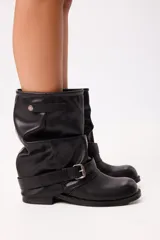 Bota de caña media estilo slouchy en color negro, confeccionada en material sintético con acabado liso. Presenta un diseño holgado en la caña, puntera redondeada, suela plana y una tira decorativa con hebilla metálica ajustable en la zona del tobillo.