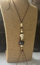 Collar largo de estilo artesanal con cordón de cuero marrón y una serie de cuentas de diversos materiales, incluyendo piedra, madera y metal dorado, dispuestas en el centro.