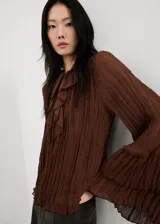Blusa de color marrón chocolate, confeccionada en tejido plisado con textura. Presenta un diseño de cuello redondo con detalle de lazo, mangas largas amplias con puños acampanados y un corte holgado con volados sutiles en la parte frontal.