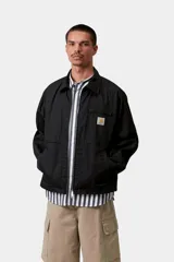 Campera Postal Jacket de Carhartt WIP en color negro, con cierre frontal de cremallera, cuello clásico, bolsillos laterales y un bolsillo con cierre en el pecho adornado con el parche del logo de la marca.