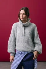 Buzo tejido en mohair de color gris, con silueta amplia y corte en línea A. Presenta cuello tipo remera con broches que permiten cerrarlo como polera, mangas largas con puños y un diseño de tejido horizontal.