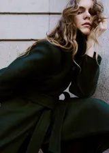 Abrigo largo de paño en color verde oscuro, con cuello alto, cierre frontal oculto y cinturón ajustable del mismo tejido. Presenta un diseño estructurado con bolsillos laterales.