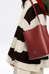 Bolso de hombro tipo cubo de estructura rígida, color marrón oscuro, confeccionado en piel vacuna. Posee asa regulable con hebilla metálica y cierre de cordón en la parte superior. Lleva el logo metálico de Bimba y Lola en la parte frontal inferior.