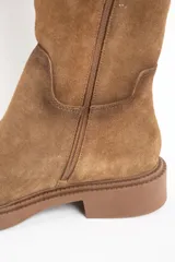 Bota de caña alta color camel, confeccionada en gamuza. Presenta una suela de goma gruesa y plana, y un diseño liso sin cierres visibles.