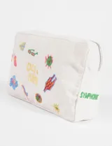 Estuche rectangular de tela color beige con cierre superior plateado. Presenta bordados coloridos con temática tropical, incluyendo texto "COCO & PIÑA" y varios íconos como gafas de sol, una flor, un traje de baño y un sol. En el lateral tiene bordado el texto "SYMPHORIE" en verde.
