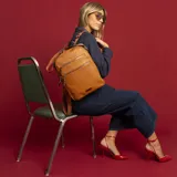 Mochila de cuero sintético con textura granulada, color verde oliva. Cuenta con dos bolsillos frontales con cierre metálico, asa superior rígida y correas ajustables de cinta textil. Incluye placa metálica decorativa en la parte inferior.