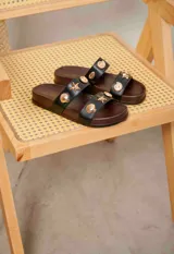 Sandalias planas de dos tiras negras con base de madera y apliques metálicos dorados en forma de caracoles y estrellas de mar.