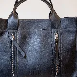 Cartera de cuero color tostado, estructurada, con doble asa de mano, correa desmontable y dos bolsillos frontales verticales con cierre. Presenta detalles de costuras pespunteadas y el logo de la marca grabado en la parte inferior.