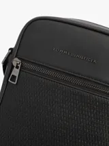 Bandolera reporter Tommy Hilfiger de color negro, confeccionada en poliuretano con acabado liso y panel frontal inferior con detalle tejido. Cuenta con un compartimento principal con cierre de cremallera, bolsillo exterior frontal también con cremallera, bolsillo interior abierto, correa ajustable y logo metálico de la marca en la parte delantera.