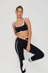 Conjunto deportivo compuesto por un top y calzas largas. El top es negro con bordes blancos y logo estampado en el frente. Las calzas son negras con cintura cruzada en V, detalles de líneas laterales blancas y logo estampado en la pierna.