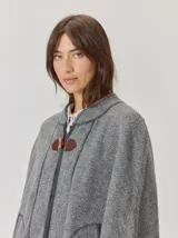 Capa de tejido de punto en color gris melange, con diseño abierto y cierre frontal mediante un botón tipo duffle de madera con detalle en cuero. Presenta bolsillos laterales amplios y bordes con costuras a la vista.