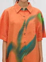 Camisa tipo vestido de corte oversize, confeccionada en viscosa. Presenta un diseño estampado con trazos abstractos en tonos verdes sobre una base naranja, cuello clásico, cierre frontal con botones y mangas cortas.