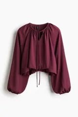 Blusa color vino tinto, con cuello redondo y escote en V con cordones para atar. Presenta mangas globo largas y un bajo fruncido con efecto burbuja.