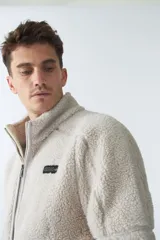 Campera de sherpa color verde militar, con cierre frontal completo, cuello alto y bolsillos laterales con cierre. Presenta un parche pequeño con logo en el pecho.