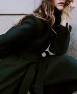Abrigo largo de paño en color verde oscuro, con cuello alto, cierre frontal oculto y cinturón ajustable del mismo tejido. Presenta un diseño estructurado con bolsillos laterales.