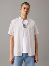 Camisa de manga corta Calvin Klein confeccionada en popelín de algodón elástico con diseño de rayas verticales en tonos beige y blanco. Presenta cuello en punta, cierre frontal de botones, bolsillo de parche en el pecho con logo bordado y bajo recto.