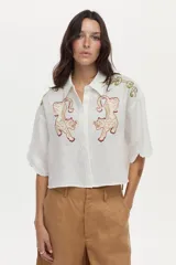 Camisa crop blanca de lino con silueta relajada, cuello clásico y mangas cortas. Presenta bordados de tigres en el frente y detalles ornamentales en los hombros.