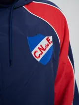 Campera deportiva Umbro del Club Nacional de Football, modelo Venti, para hombre. Diseño en color blanco con mangas azules y detalles de líneas rojas. Cuenta con capucha con cordón ajustable, cierre frontal y bolsillos. Presenta el logo de Umbro y el escudo del club bordados en el pecho.