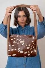 Cartera de hombro con diseño de animal print tipo vaca en tonos marrón y blanco. Presenta una correa ancha de gamuza marrón y una placa metálica con logo en el frente.