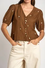 Blusa de viscosa color marrón, con escote en V, mangas cortas abullonadas y cierre frontal con botones. Cuenta con pequeños detalles bordados distribuidos por la prenda.