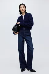 Pantalón de jean estilo wide leg con tiro alto, confeccionado en denim de algodón con leve stretch. Presenta cierre frontal con doble botón metálico, pliegues frontales y traseros, y bolsillos traseros de parche.