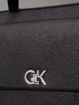 Cartera tipo shopper mediana de Calvin Klein, color negro, confeccionada en material textil con textura de lino tejido. Presenta asas fijas para el hombro, cierre de cremallera superior, bolsillo exterior frontal con cierre giratorio y herraje con monograma CK.