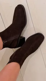 Bota de caña alta de gamuza en color marrón chocolate, con taco cuadrado y puntera cuadrada. Cuenta con un diseño holgado en la caña y detalle de trabilla decorativa en el tobillo.