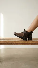 Bota corta de cuero color marrón, con diseño que combina cuero liso y sectores con textura grabada. Presenta una base robusta con taco medio y cierre lateral para facilitar el calce.