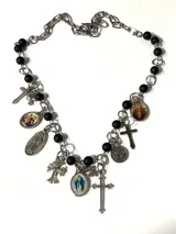 Collar tipo rosario con cadena de acero quirúrgico y perlas de vidrio esmaltadas en color negro mate. Presenta múltiples dijes colgantes de temática religiosa, incluyendo cruces, medallas con imágenes de santos y la Virgen.