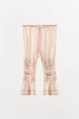 Pantalón de tiro medio confeccionado en viscosa, con cintura elástica ajustable mediante lazada. Presenta detalles de pliegues, bordados florales a tono y aplicaciones de encaje a contraste en la parte inferior, finalizando con un bajo acabado en volantes.