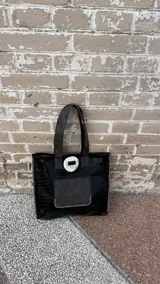 Tote bag de ecocuero negro con textura croco, cuenta con un bolsillo frontal de gamuza marrón y un detalle decorativo circular de guampa en el cierre superior. Posee doble asa de hombro y costuras visibles en los bordes.
