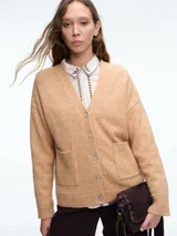 Cardigan de punto color beige con cierre frontal de botones, cuello en V y dos bolsillos delanteros. Presenta mangas largas con puños acanalados y un diseño de corte holgado.