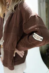 Cardigan de punto color marrón, abierto con botones imitación hueso, manga larga con parche bordado y diseño texturado.
