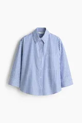 Camisa corta oversize de popelina de algodón con rayas verticales blancas y azules, cuello clásico, cierre frontal de botones, bolsillo en el pecho y mangas tres cuartos con puños anchos.